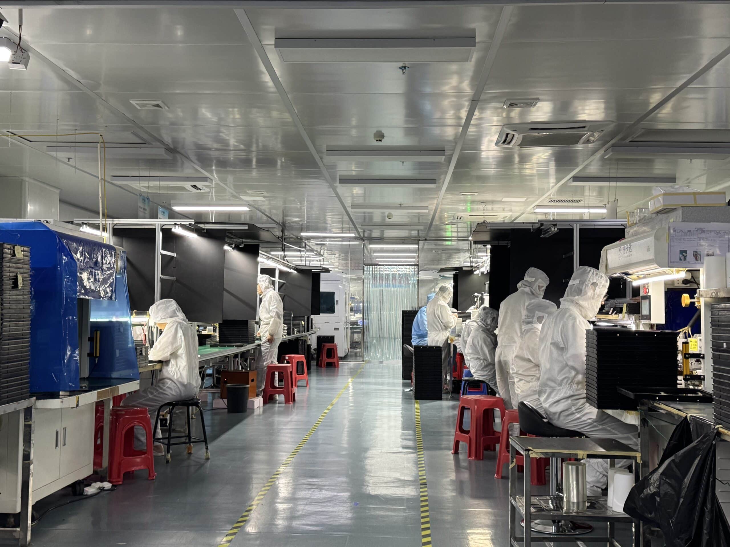 Cleanroom electronics assembly line with workers in white protective suits at workstations, red stools, yellow-black floor markings, and overhead lighting.